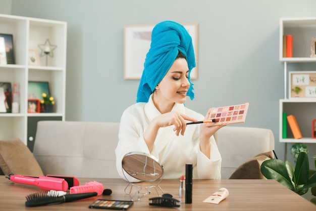 Молодая женщина, держащая и смотрящая на палитру теней с кисточкой для макияжа, завернула волосы в полотенце, сидя за столом с инструментами для макияжа в гостиной