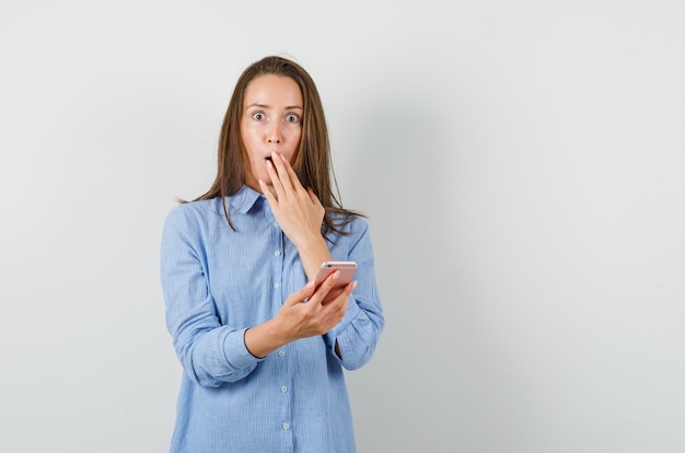 Young woman holding mobile phone in blue shirt and looking shocked.