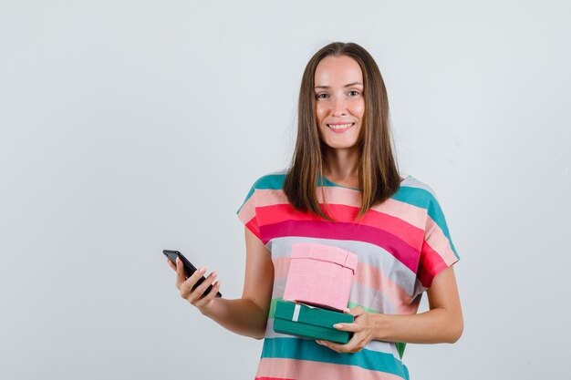 Молодая женщина держит мобильный телефон и подарочные коробки в футболке и рад, вид спереди.