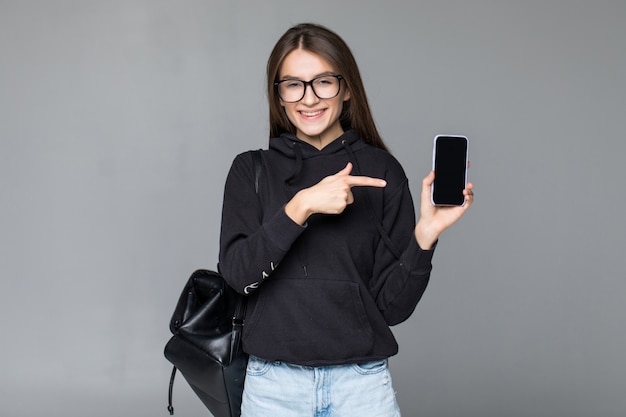 Young woman holding a mobile phone and pointing at it isolated on white wall