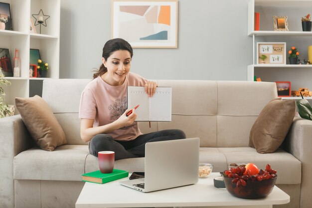 リビングルームでラップトップを見ているコーヒーテーブルの後ろのソファに座ってノートを保持している若い女性