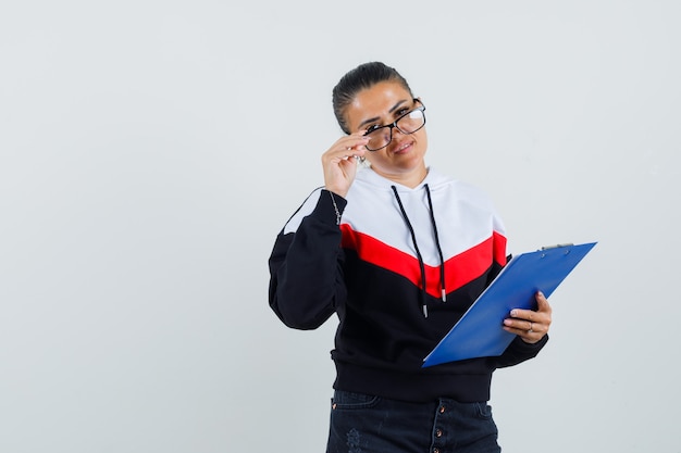 Giovane donna che tiene il taccuino mentre si toglie gli occhiali in maglione, jeans neri e sembra carina. vista frontale.