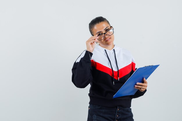 Young woman holding notebook while taking off glasses in sweater, black jeans and looking pretty. front view.