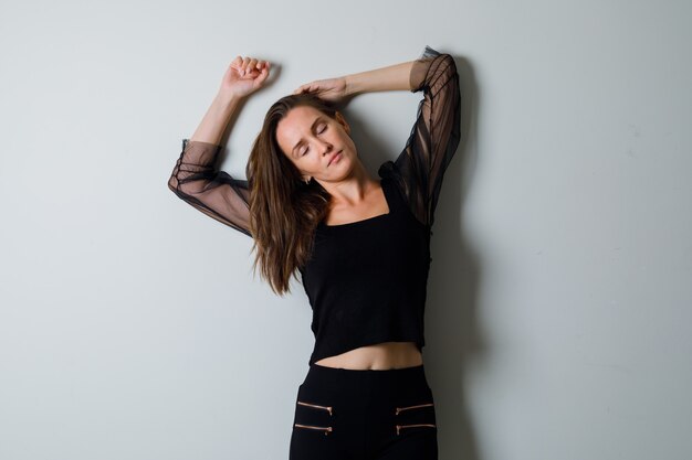 Young woman holding one arm over head, raising another arm and closing eyes in black blouse and black pants and looking charming