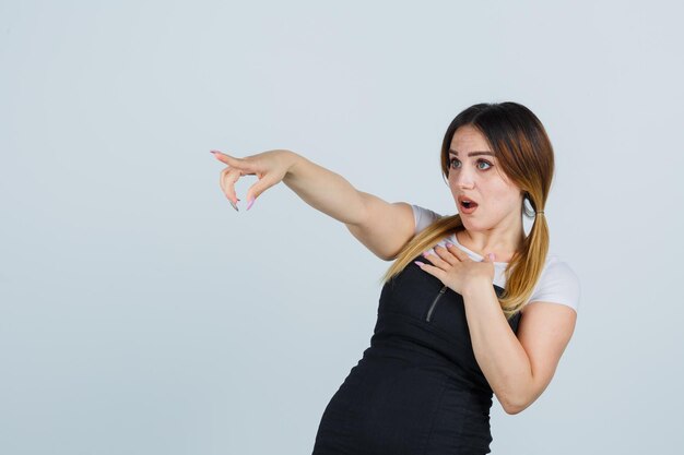 Young woman holding one hand over chest while pointing away and looking surprised