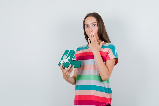 Young woman holding opened gift box in t-shirt and looking amazed. front view.