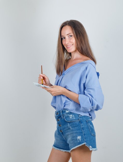 Young woman holding paint brush over palette in shirt, shorts and looking cheerful.