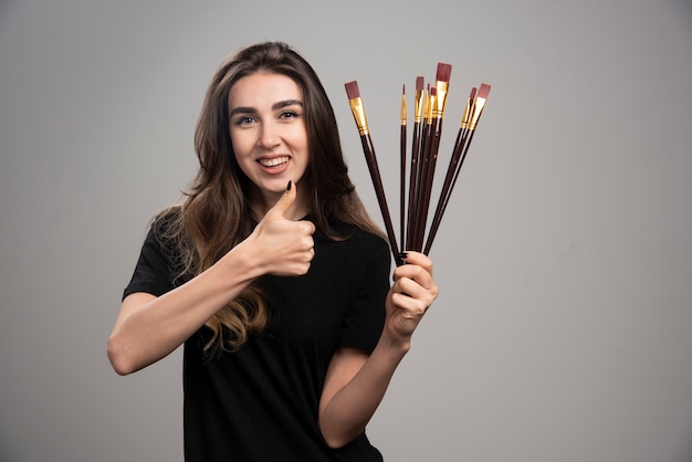 Young woman holding paint brushes and giving thumbs up. 