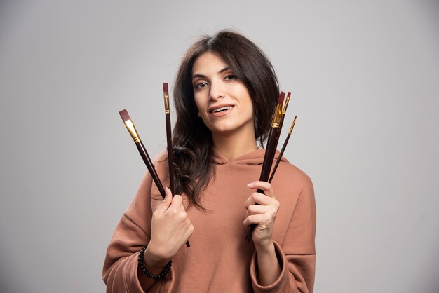 Young woman holding paint brushes on gray