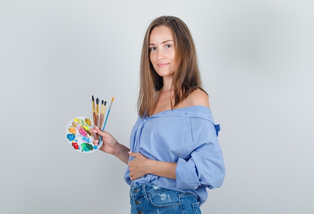 Giovane donna che tiene gli strumenti di pittura in camicia blu, pantaloncini e guardando fiducioso.