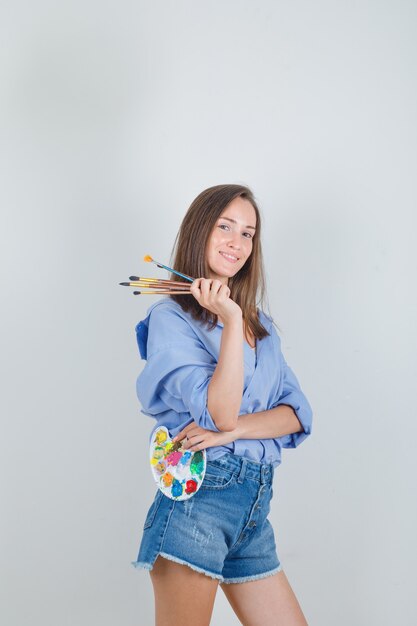 Молодая женщина, держащая инструменты для рисования в голубой рубашке, шортах и выглядящая счастливой.