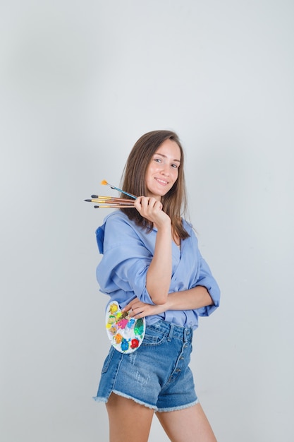 Young woman holding painting tools in blue shirt, shorts and looking happy