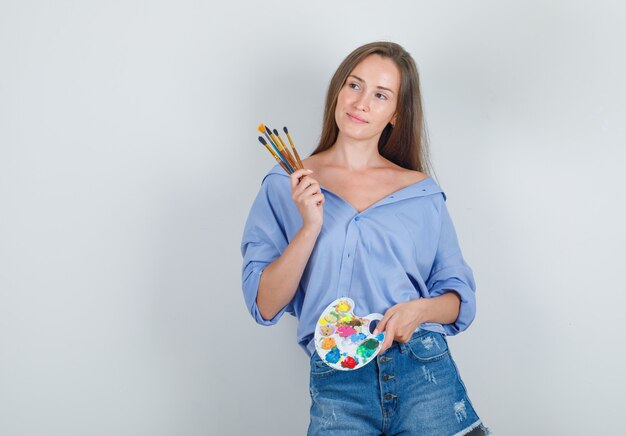 Young woman holding painting tools in shirt, shorts and looking glad