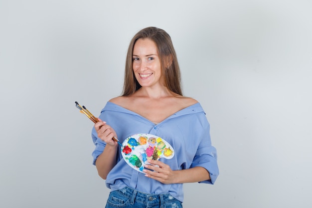 Young woman holding painting tools in shirt, shorts and looking jolly