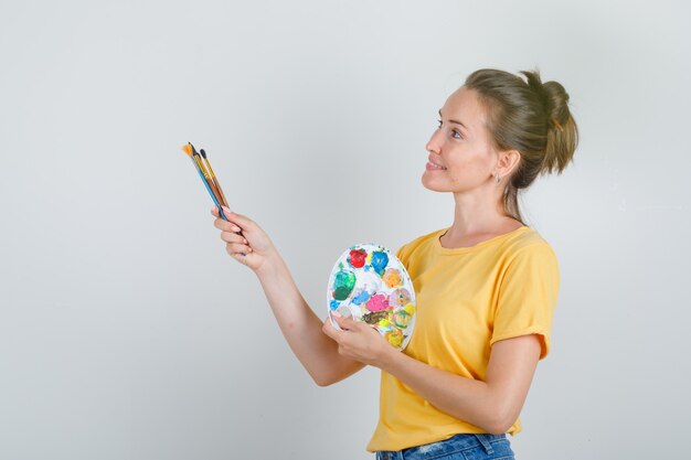 Молодая женщина, держащая инструменты для рисования, глядя вверх в желтой футболке, джинсовых шортах и выглядит веселой.