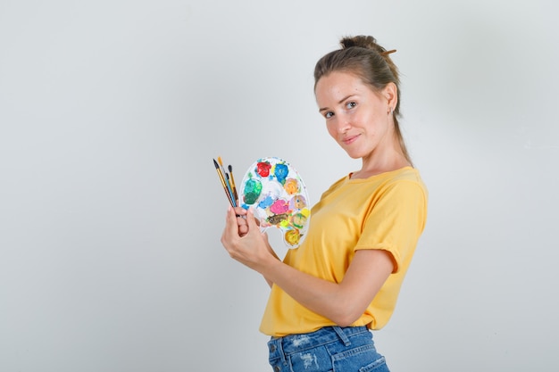 Young woman holding painting tools in yellow t-shirt, jeans shorts and looking cheerful .