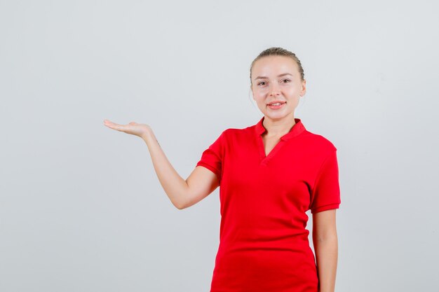Young woman holding palm to show something in red t-shirt and looking cheerful