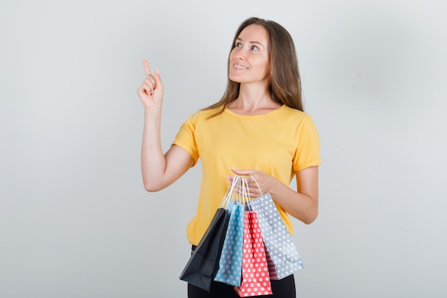 young woman holding paper bags with finger up in yellow t-shirt, black pants and looking glad