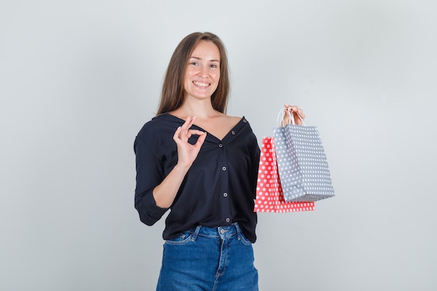 Giovane donna che tiene i sacchetti di carta con il segno giusto in camicia nera, pantaloncini di jeans e che sembra felice