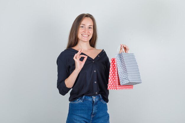 Молодая женщина держит бумажные пакеты с знаком ОК в черной рубашке, джинсовых шортах и выглядит счастливой