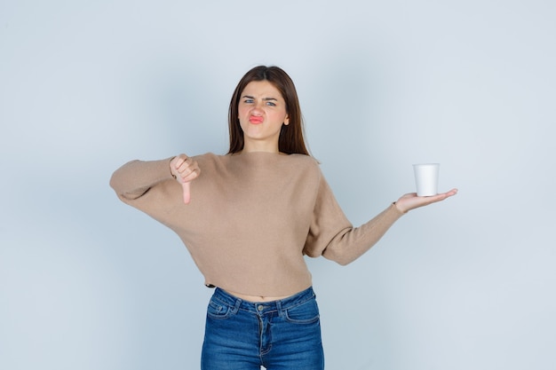 Young woman holding paper cup, showing thumb down in sweater, jeans and looking displeased , front view.