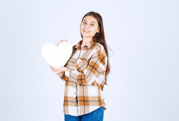 Young woman holding paper heart-shaped card on white wall. 