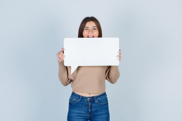 Young woman holding paper speech bubble in sweater, jeans and looking happy, front view.