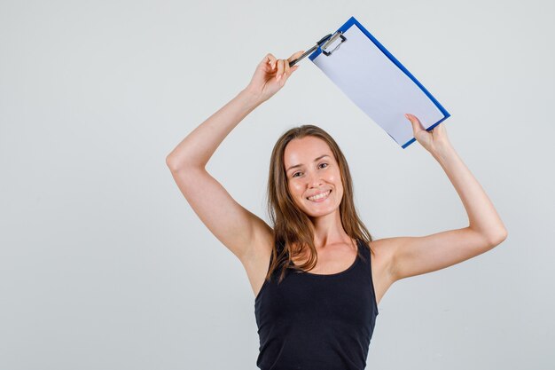 Young woman holding pen and clipboard over head in singlet and looking glad