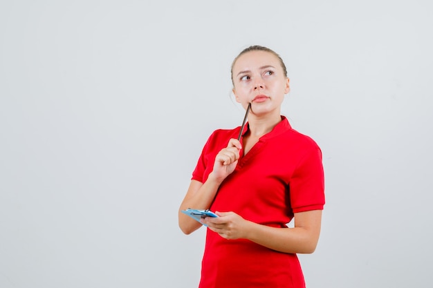 赤いTシャツに鉛筆とクリップボードを保持し、物思いにふける若い女性