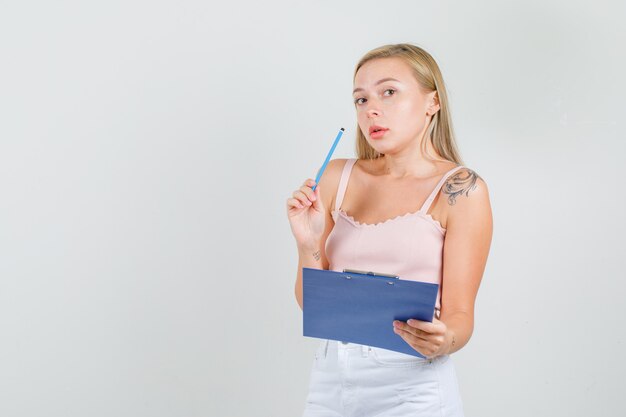 Young woman holding pencil and clipboard in singlet, mini skirt and looking busy