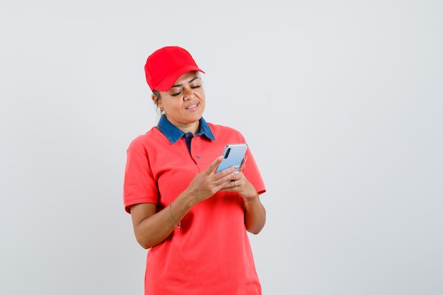 Young woman holding phone and writing text in red shirt and cap and looking focused , front view.