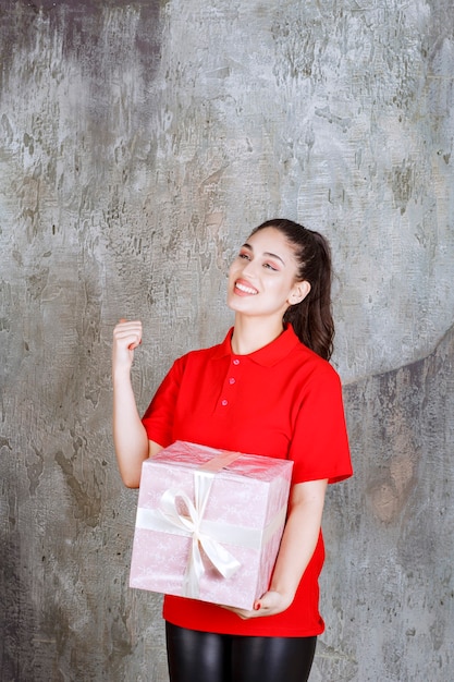 Young woman holding a pink gift box wrapped with white ribbon and showing positive hand sign.