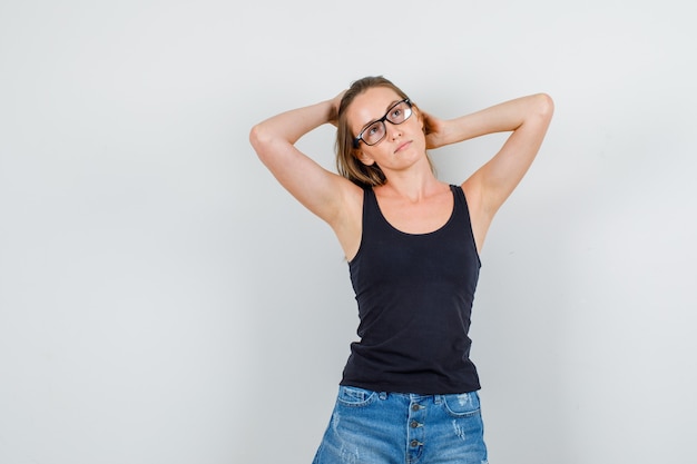 Young woman holding raised hands on hair in singlet, shorts, glasses and looking pensive