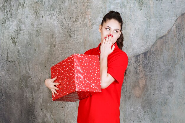 Молодая женщина, держащая красную подарочную коробку с белыми точками на ней, выглядит задумчивой.