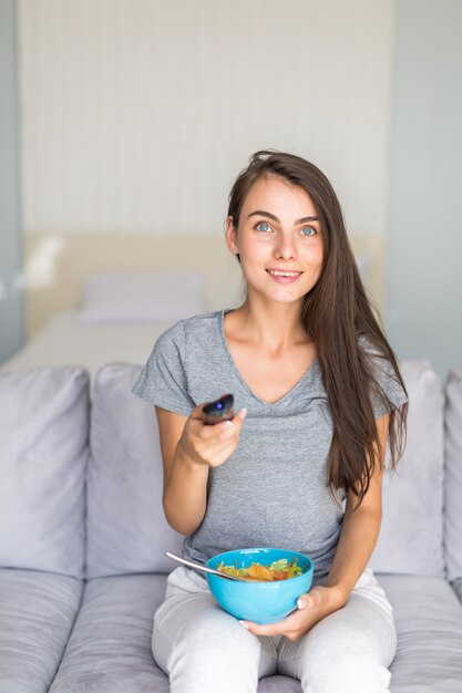 Молодая женщина, держащая пульт дистанционного управления дома, сидя на диване и смотреть телевизор с салатом