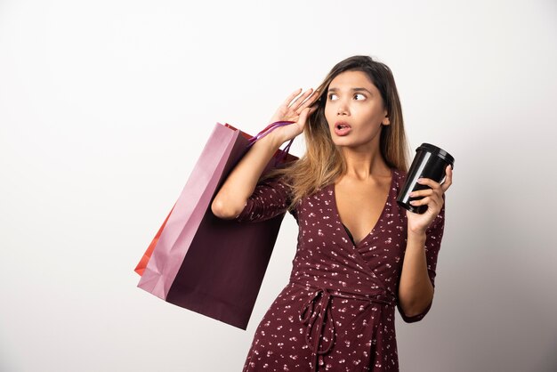 Young woman holding shop bags and a cup of drink on white wall. 