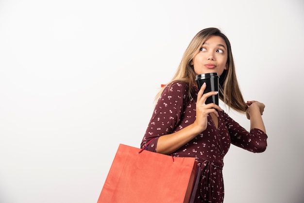 Young woman holding shop bags and a cup of drink on white wall. 