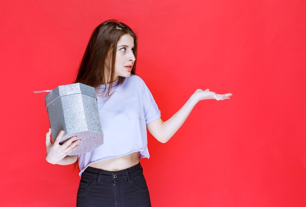 Free photo young woman holding a silver gift box and pointing at someone.