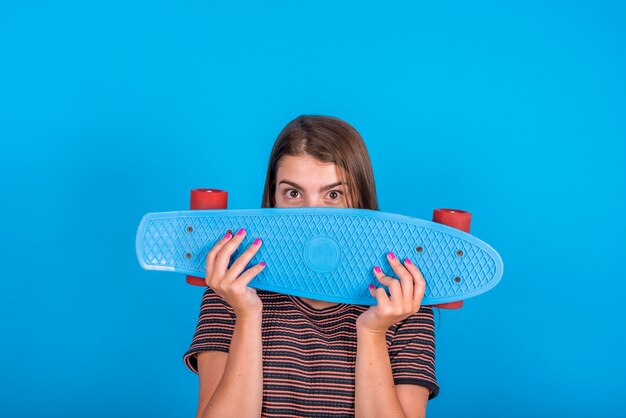 青の背景に顔の前でスケートボードを保持している若い女性