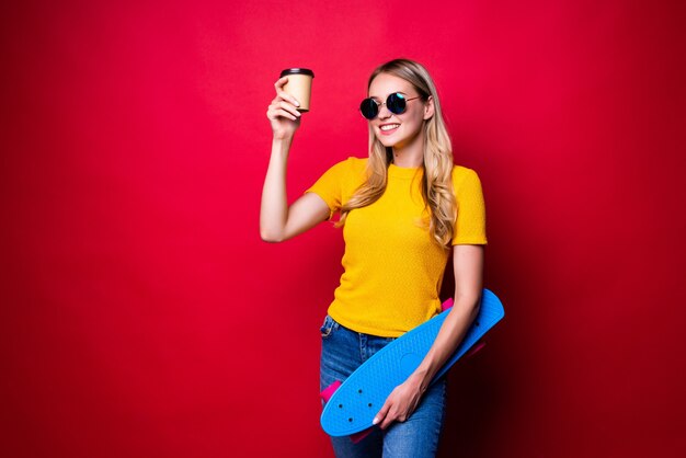 肩にスケートボードと赤い壁に対して隔離のコーヒーを保持している若い女性