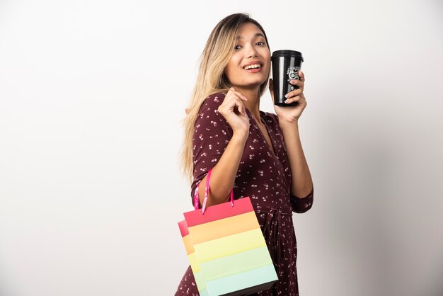 Young woman holding a small shop bag and a cup of drink on white wall.