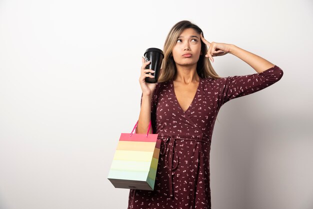 Young woman holding a small shop bag and a cup of drink on white wall.