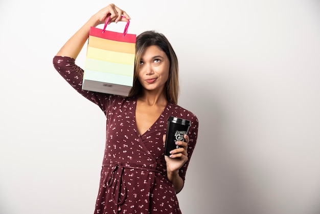 Young woman holding a small shop bag and a cup of drink on white wall. 