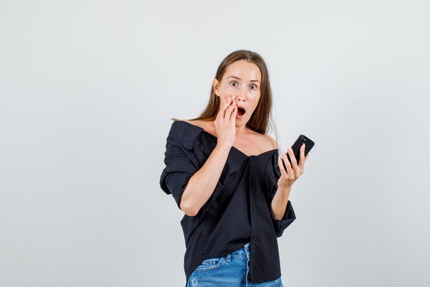 Young woman holding smartphone in shirt, shorts and looking surprised