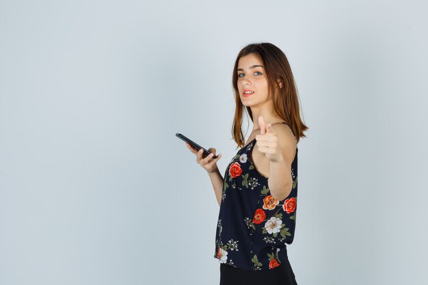 Young woman holding smartphone, showing thumb up in blouse, skirt and looking confident .