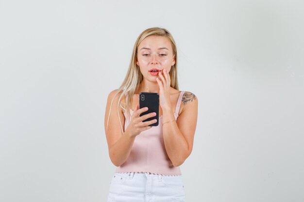 Young woman holding smartphone in singlet, mini skirt and looking surprised.