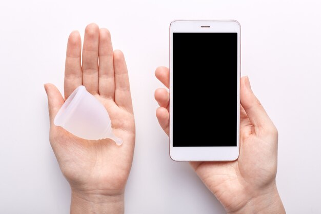 Young woman holding smartphone with blank screen in one hand and white clean menstrual cup