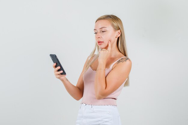 Young woman holding smartphone with finger on cheek in singlet