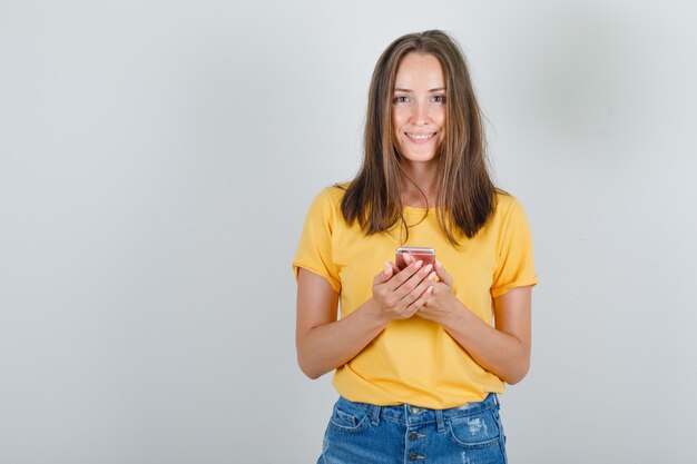 Young woman holding smartphone in yellow t-shirt, shorts and looking cheerful