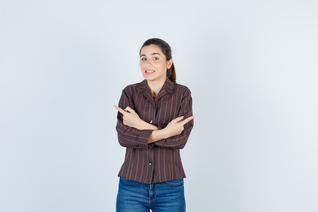 Young woman holding two arms crossed, pointing opposite directions with index fingers in striped shirt, jeans and looking happy , front view.